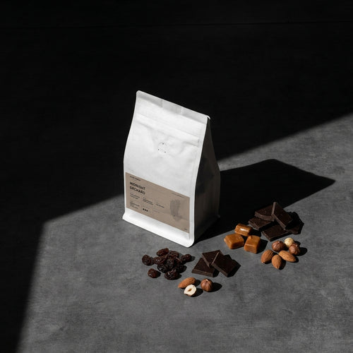 White coffee bag with brown label on a dark surface with chocolate pieces, caramel, raisin and nuts.